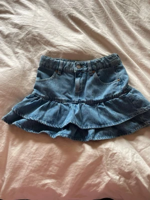 Blå volangkjol i denim - Söt kort jeanskjol med volangkant och klassiska fickor fram. Kjolen har bälteshällor och stängs med knapp och dragkedja. Perfekt för en avslappnad och trendig look.