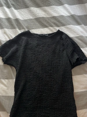 Svart ribbad topp med kort ärm - Säljer en svart ribbad topp med kort ärm och rund hals. Tyget har en stretchig, strukturerad känsla som ger en cool look. Perfekt att styla med jeans eller kjol för en trendig outfit.