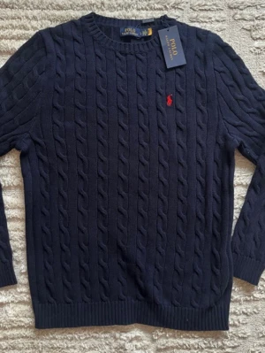 Mörkblå kabelstickad tröja Polo Ralph Lauren - Kabelstickad mörkblå tröja från Polo Ralph Lauren med rund halsringning och den klassiska röda broderade logotypen på bröstet. Tröjan har ribbade muddar vid ärmslut och nederkant. Perfekt för en stilren och tidlös look.