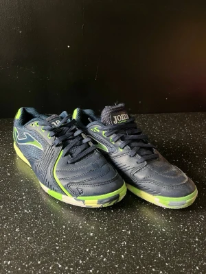 Joma blå-gröna inomhusfotbollsskor - Snygga inomhusfotbollsskor från Joma i marinblått med gröna detaljer och vit logga på sidan. Skorna har snörning, mönstrad sula i grönt, svart och vitt för bra grepp på planen. Perfekta för futsal eller inomhusträning.