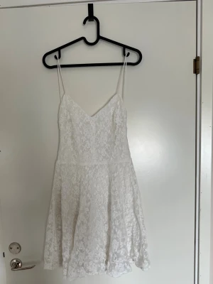 Vit spetsklänning med tunna axelband - Kort vit klänning med tunna axelband och v-ringning, täckt av vacker spets med blommönster. Perfekt för skolavslutning eller studenten!