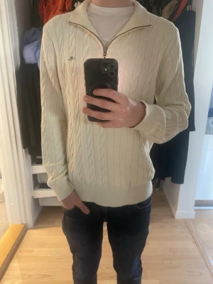 Vit kabelstickad tröja Gant - Kabelstickad vit tröja från Gant med halv dragkedja och hög krage. Tröjan har ribbade muddar vid ärmslut och nederkant med Gant-logga på bröstet. Ändast använd fåtals gånger. 
