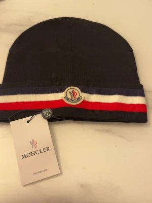Svart Moncler mössa med logga - Snygg svart mössa från Moncler med klassisk Moncler-logga framtill. Mössan har en uppvikt kant med randiga detaljer i blått, vitt och rött. Tillverkad i mjukt stickat material som håller dig varm och stilren under kalla dagar.