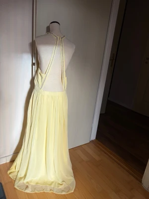 Ljusgul långklänning med öppen rygg - Elegant ljusgul långklänning med öppen rygg och tunna axelband. Klänningen har ett luftigt, svepande fall och är tillverkad i ett lätt, skirt material. 