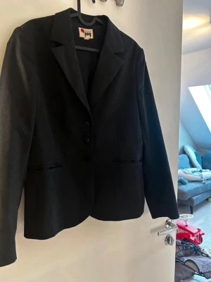 Svart kavaj från Joy med smal passform - Stilren svart kavaj från Joy med tre knappar framtill och smal passform. Klassisk krage och två fickor framtill. Materialet ser ut att vara en blandning med diskret struktur, perfekt för en snygg och enkel look. Innertyget på ena fickan har gått sönder men utöver det inget fel på den. Rök och djurfritt hem. Pruta gärna!🌸