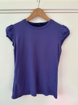 Lila topp med puffärmar - French Connection - storlek S - En söt lila topp från French Connection i storlek S med puffade korta ärmar och rund halsringning. Toppen har en slim passform och är tillverkad i mjuk bomull, perfekt för en trendig och bekväm look. Fint skick!