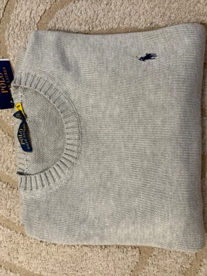 Ljusgrå stickad tröja Polo Ralph Lauren - Snygg ljusgrå stickad tröja från Polo Ralph Lauren med klassisk rund halsringning och ribbade muddar. Tröjan har den ikoniska mörkblå logotypen broderad på bröstet. Perfekt för lager på lager under vintern.