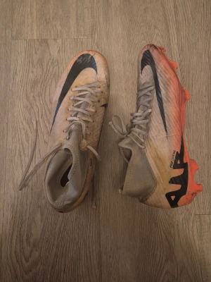 Nike Mercurial fotbollsskor råsa - Säljer ett par Nike Mercurial fotbollsskor med högt skaft och snörning. Skorna är ljusbeige med svart Nike-logga och har en råsa sula med dobbar. Materialet är syntet och designen är aerodynamisk för snabbhet på planen.