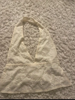 Beige broderad halterneck topp - Säljer en beige halterneck topp med broderade blommor och knytband i ryggen. Toppen har en v-ringad front och är ärmlös, perfekt för varma dagar. Materialet känns lätt och luftigt, och broderierna ger en romantisk touch.
