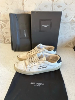Saint Laurent vita sneakers - Snygga vita sneakers från Saint Laurent med svart hälparti och handskriven logga på sidan. Skorna har klassisk låg profil, beige sula och är tillverkade i mocka och läder. Perfekta för dig som gillar exklusiva och trendiga märken.