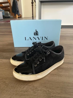 Svarta Lanvin sneakers med lackdetaljer - Snygga svarta sneakers från Lanvin med ovandel i mocka och glansiga lackdetaljer på tån. Skorna har vita sulor och svarta skosnören. Klassisk låg modell som passar till många olika stilar. Kommer med originalkartong och dustbag. Förkommer lite slitningar i mockan men övrigt bra skick :)storlek uk6 passar ca42.  skriv vid frågor eller om du vill diskutera priset!