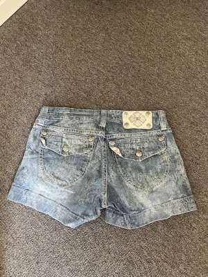 Blå denimshorts från Miss Me - Snygga blå denimshorts från Miss Me med blommiga detaljer på insidan av linningen och dekorativa knappar på bakfickorna. Shortsen har en klassisk femficksdesign och coolt tvättad look. Perfekta för varma dagar och enkel att matcha med allt.