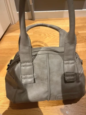 Grå handväska i mjukt material - Snygg grå handväska med två rejäla handtag och stilren design. Väskan är tillverkad i ett mjukt, matt material och har dekorativa sömmar framtill. Perfekt storlek för att bära med sig det viktigaste och passar till många olika outfits.