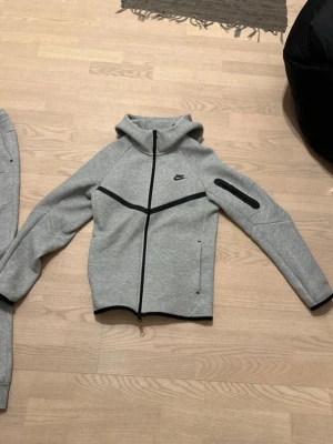 Nike tech fleece set - Snyggt grått loungewear set från Nike med zip-hoodie och matchande byxor. Hoodien har svarta detaljer, dragkedja och fickor, samt en extra ficka på ärmen. Byxorna har dragsko i midjan och muddar vid bensluten. Perfekt för chill dagar hemma.
