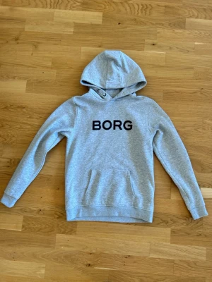 Grå hoodie från BORG - Snygg grå hoodie från BORG med svart tryck på bröstet. Klassisk modell med huva och känguruficka framtill. Tillverkad i mjukt sweatshirtmaterial som är skönt mot huden. Perfekt för en avslappnad och sportig stil.