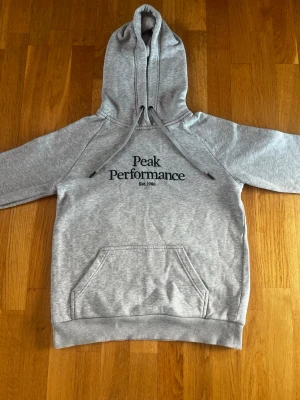 Grå hoodie Peak Performance - Snygg grå hoodie från Peak Performance med svart logga på bröstet och ficka framtill. Klassisk modell med huva. Tillverkad i mjukt sweatshirtmaterial som är perfekt för vårdagar.   Hoodien är i mycket bra skick och har inga fläckar eller skador. Nypris 1300kr och den är använd ett fåtal gånger! Hör av er vid funderingar! 