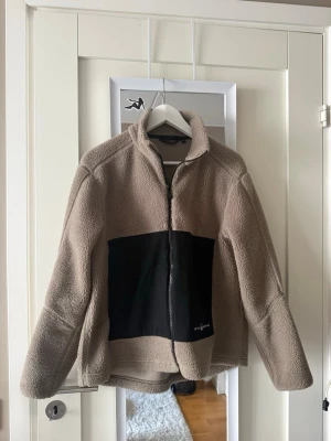 Beige teddyjacka från sail racing  - Mysig teddyjacka i beige med svart panel framtill och dragkedja. Jackan har hög krage och är tillverkad i mjukt, fluffigt material som håller dig varm. Perfekt för att skapa en chill och trendig look under kalla dagar.