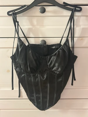 Svart korsett med smala axelband  - Stilren svart korsett med smala justerbara axelband och markerade sömmar. Korsetten har en glänsande finish och dragkedja bak för enkel på- och avtagning. Materialet är syntetiskt och ger en cool look. 🖤