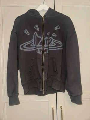 Svart hoodie från Broken Planet - Cool svart hoodie från Broken Planet med stort grafiskt tryck på framsidan och text på huvan. Tröjan har dragkedja, ribbade muddar och en stor ficka framtill. Perfekt för dig som gillar streetwear och vill sticka ut med unik design.