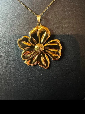 Guldigt halsband med stor blomma - Unikt halsband med ett stort, blommigt hänge i guldfärgad metall. Hänget har en detaljerad blomform med markerade kronblad och en texturerad mitt. Kedjan är tunn och matchar hängets färg. Perfekt för dig som vill sticka ut med ett statement-smycke.