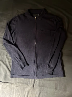 Mörkblå ziptröja från J.Lindeberg - Mörkblå ziptröja från J.Lindeberg med krage och bröstficka. Tröjan har långa ärmar, ribbade muddar och är tillverkad i ett mjukt stickat material. Perfekt för lager-på-lager och enkel att matcha med jeans. Det står att den är L men är mer som S-M