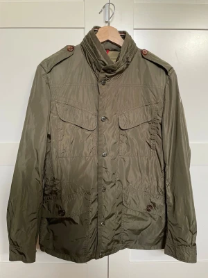 Grön moncler fieldjacka  - Tja! Säljer en snygg lite äldre grön fieldjacka med hög krage, fyra framfickor med snygga knappar samt axelklaffar. Jackan är rätt så tunn vilket gör den perfekt nu till våren. Den går att knäppa åt vid ländryggen men även tighta till.