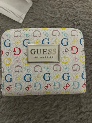 Vit plånbok från Guess med färgglatt mönster - Snygg vit plånbok från Guess med färgglada G mönster i blått, rött och gult. Plånboken har en metallplatta med Guess-loggan på framsidan och är tillverkad i ett strukturerat syntetmaterial. Perfekt för att hålla koll på kort och cash. Jag fick den i present och är lite osäker på om den är äkta