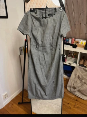  klänning  - Stilren klänning med diskret rutigt mönster och korta ärmar. Klänningen har dekorativa knappar vid halsringningen och markerad midja. Perfekt för dig som gillar klassisk och tidlös stil. Klänningen är svart och vit.