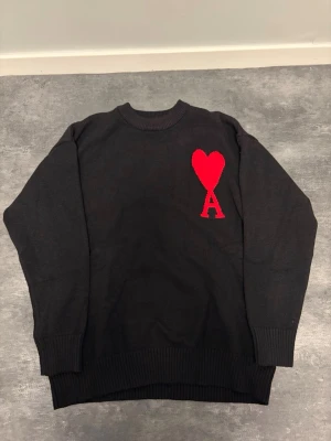 Svart stickad tröja från Ami Paris - Svart stickad tröja från Ami Paris med röd hjärtlogga och bokstaven A på bröstet. Tröjan har rund halsringning, långa ärmar och ribbstickade muddar vid ärmslut och nederkant. Perfekt för en stilren och trendig look.