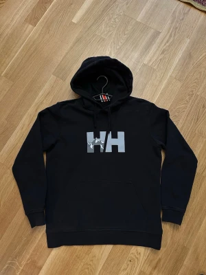 Svart hoodie från Helly Hansen - Svart hoodie från Helly Hansen med stor HH-logga i grått på bröstet. Klassisk passform, med luva och stor magficka. Perfekt för dig som gillar streetwear och enkel stil. Materialet är mjukt och bekvämt