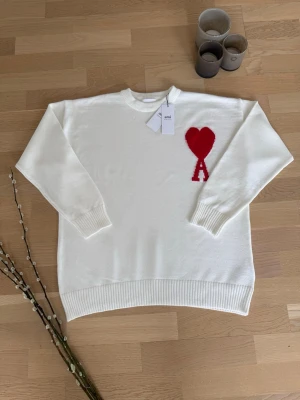 Vit stickad tröja Ami Paris - Vit stickad tröja från Ami Paris med röd hjärtlogga och bokstaven A på bröstet. Tröjan har rund halsringning, ribbade muddar och nederkant. Tillverkad i mjukt material som ger en clean och stilren look.