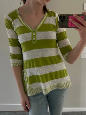 Grön och vit randig v-ringad topp - Grön och vit randig topp med v-ringning och trekvartsärm. Toppen har knappar vid halsen och en avslappnad passform. Tillverkad i mjukt bomullsmaterial, perfekt för en avslappnad stil.