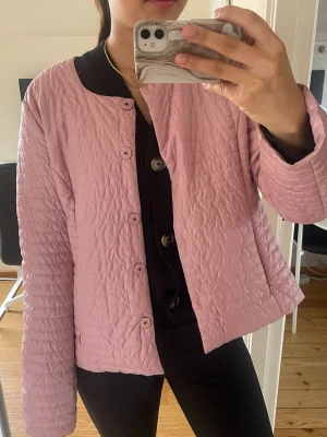 Ljusrosa quiltad cardiganjacka - Säljer en ljusrosa quiltad cardiganjacka med rund halsringning och knäppning framtill. Jackan har en lätt glansig yta och raka ärmar. Perfekt för lager på lager och ger en chill men ändå stylish vibe.