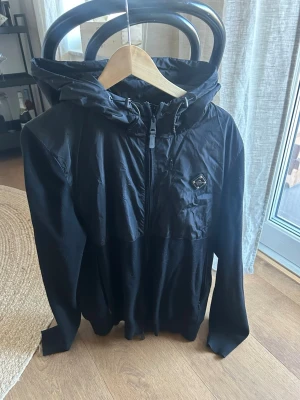 Svart vindjacka med huva - Svart vindjacka med huva och half zip framtill. Jackan har en modern look med en liten logga på bröstet och är tillverkad i syntetmaterial. Perfekt för blåsiga dagar och enkel att matcha med olika outfits. Jackan är helt felfri på utsidan men i höger innerficka är det ett litet hål. Annars väldigt bra skick.