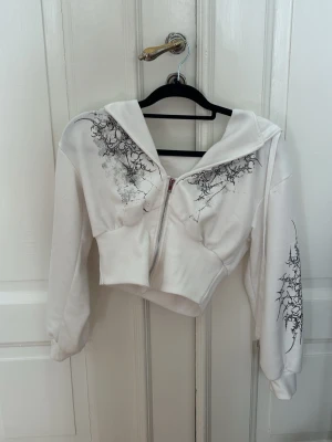 Vit cropped hoodie med tribaltryck - Säljer en vit cropped hoodie med dragkedja framtill och svart tribalinspirerat tryck på bröstet och ärmarna. Hoodien har huva med dragsko och ribbade muddar. Perfekt för en edgy streetwear-look. Materialet känns mjukt och bekvämt.