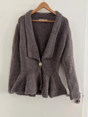 Rabens Saloner Hand-Knitted Alpaca Blend Peplum Cardigan – Size S/M - Beautiful, chunky hand-knitted cardigan from the boutique brand Rabens Saloner. This piece features a flattering peplum waist, a classic shawl collar, and a stunning decorative silver-tone rose button. • Material: Wool/alpaca blend  • Fit: Fits like a Size S or a small M. The peplum silhouette creates a very feminine shape. • Condition: Good pre-loved condition. • Color: Neutral taupe / dark grey melange. Perfect for layering.