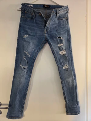 Ljusblå slitna jeans Jack & Jones - Säljer ett par ljusblå jeans från Jack & Jones med flera slitningar och hål på benen för en trendig look. Jeansen har klassisk femficksmodell, knappgylf och smal passform. Perfekta för dig som gillar en avslappnad och modern stil.