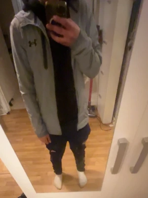 Under Armour jacka - Snygg ljusgrå vindjacka från Under Armour med dragkedja och huva. Jackan har en minimalistisk design med logga på bröstet och smala muddar vid ärmsluten. Perfekt för dig som vill ha en sportig och stilren look.