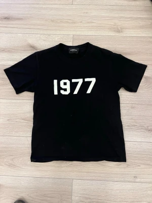 Essentials t-shirt 1977 - Svart t-shirt från Essentials med vit text '1977' tryckt på bröstet. Klassisk passform med korta ärmar och rund hals. Perfekt för dig som gillar stilrena och trendiga plagg med en retrovibe.