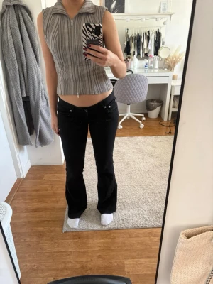 Svarta bootcut jeans med låg midja - Säljer ett par svarta bootcut jeans med låg midja. Jeansen har en figurnära passform upptill och lätt utsvängda ben. Snygga detaljer med silverknappar och bälteshällor. Perfekta för en trendig och avslappnad look.