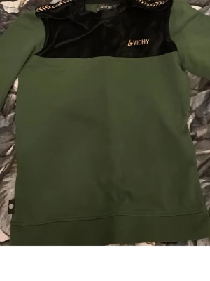 Grön hoodie från Vichy med sammet - Stilren grön hoodie från Vichy med svart sammetsdetalj över bröstet och axlarna. Broderad Vichy-logga i guld på vänster bröst. Klassisk passform och rund halsringning. Perfekt för dig som vill ha en unik och trendig look.