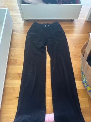 Svarta mjukisbyxor i velour - Säljer ett par svarta mjukisbyxor i mjuk velour med raka ben och dragsko i midjan. Byxorna har fickor bak med knappdetaljer och är perfekta för en chill och avslappnad stil. Passar dig som gillar bekväma och snygga plagg.