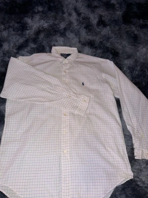 Vit rutig skjorta från Polo Ralph Lauren - Säljer en klassisk vit skjorta från Polo Ralph Lauren med rutigt mönster i blått och gult. Skjortan har lång ärm, knappar framtill och en broderad logga på bröstet. Perfekt för dig som gillar stilrena plagg med snygga detaljer.