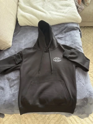 Svart hoodie från Amiri med tryck - Svart hoodie från Amiri med vit logga och texttryck både fram och bak. Klassisk känguruficka framtill, ribbade muddar och justerbar huva med snören. Tillverkad i mjukt bomullsmaterial för en skön och avslappnad look. Priset kan diskuteras!