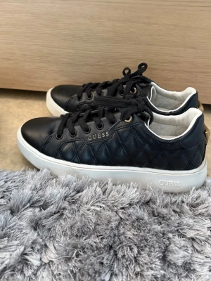 Svarta Guess sneakers med vit sula - Stilrena svarta sneakers från Guess med quiltad läderlook och vit sula. Skorna har svarta snören, guldfärgade detaljer och Guess-logga på sidan och sulan. Perfekta för en trendig och avslappnad stil. 