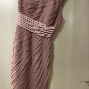 Ljusrosa one shoulder klänning från Pronovias - Elegant ljusrosa klänning från Pronovias med one shoulder-design och draperad midja. Klänningen har ett vackert vågformat mönster i tyget och markerad midja med breda band. Perfekt för bal eller fest.