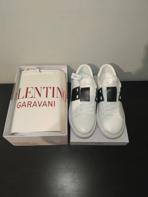 Valentino Garavani vita sneakers - Snygga vita sneakers från Valentino Garavani med svart band och nitar på sidorna. Skorna har en klassisk låg siluett, rund tå och platt vit sula. Tillverkade i läder med märkets logga på plösen. Perfekta för en trendig och stilren look.