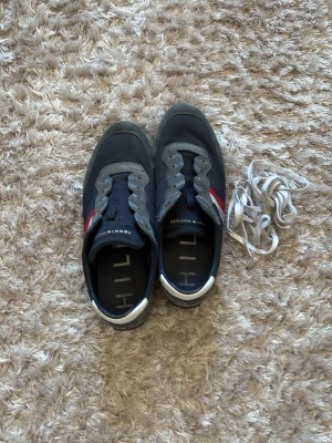 Tommy Hilfiger blå låga sneakers - Snygga blå sneakers från Tommy Hilfiger med mesh och mockadetaljer. Skorna har klassiska röda och vita ränder på sidan och vit häl. Låg modell med vita skosnören som medföljer. Perfekta för en sportig och avslappnad stil.