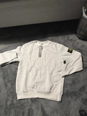 Grå sweatshirt från Stone Island - Snygg ljusgrå sweatshirt från Stone Island med klassisk rund hals och ribbade muddar. Tröjan har den ikoniska Stone Island-loggan på vänster ärm och är tillverkad i mjukt bomullsmaterial. Perfekt för en avslappnad och trendig stil.
