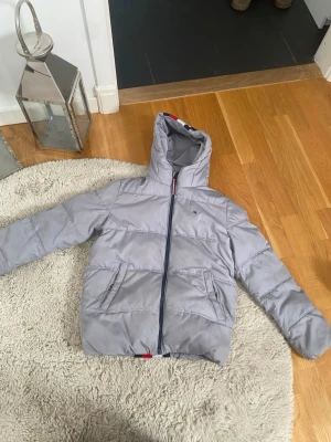 Ljusgrå pufferjacka Tommy Hilfiger - Snygg ljusgrå pufferjacka från Tommy Hilfiger med huva och dragkedja framtill. Jackan har två stora fickor med lock och detaljer i rött, vitt och blått på huva och nederkant. Perfekt för kalla dagar och riktigt skön att bära. Priset kan diskuteras 